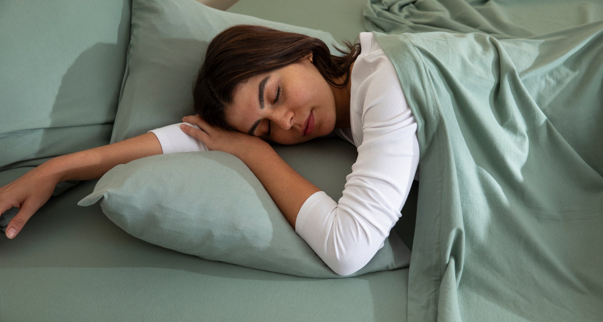 Bed sheets in jersey fabric by It-Fits, featuring a model peacefully sleeping on a sage-coloured fitted sheet and pillowcase, covered by a breathable flat sheet.