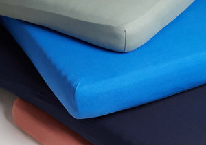 Fitted Sheets made from Cotton Jersey Fabric in green, blue, and pink randomly stacked on top of each other showcasing their snug, wrinkle-free fit and smooth material