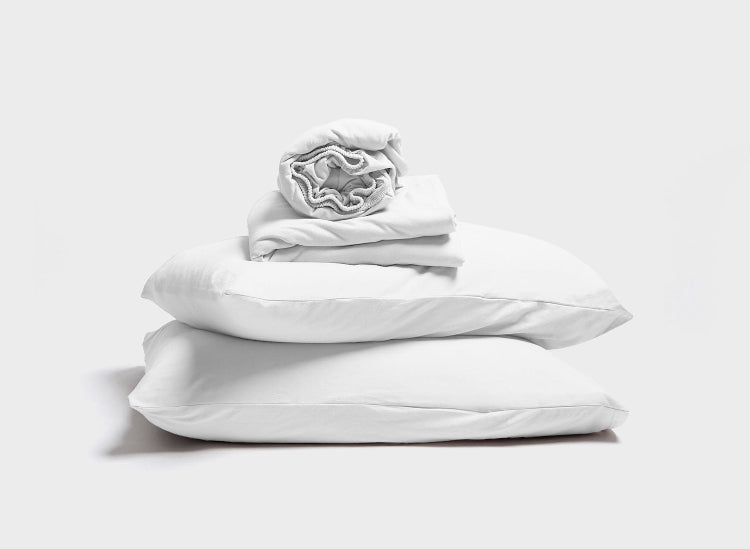 White adjustable bed sheet set by It-Fits, featuring a stack of pillows with pillowcases, along with a folded deep pocket fitted sheet and a flat sheet